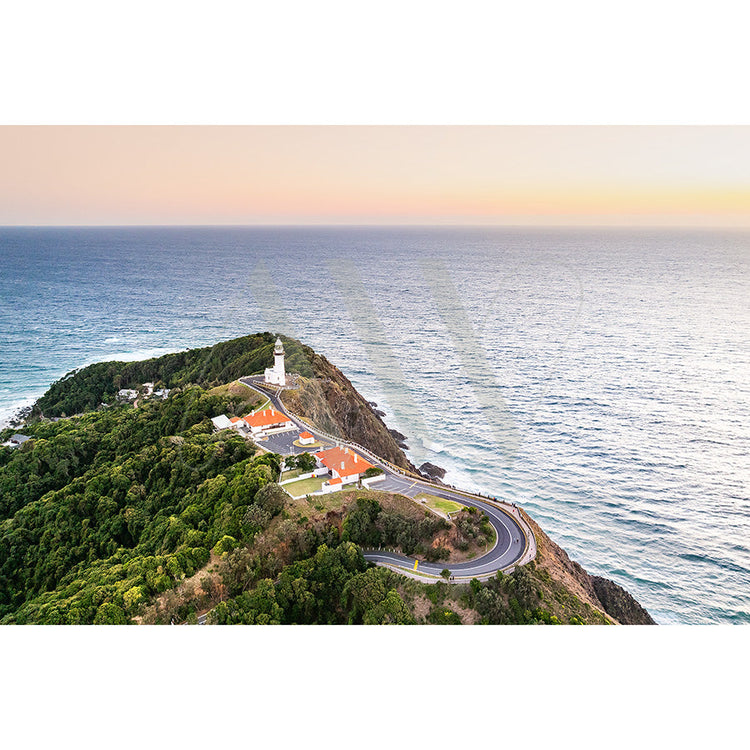 Aerial view of a lighthouse on a rocky coastline surrounded by lush greenery, with a winding road leading to it. The ocean stretches to the horizon under a pastel-colored sky.