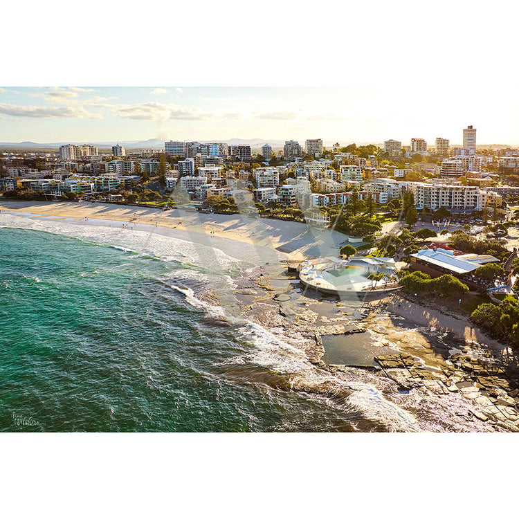 Aerial view of a coastal city with sandy beaches, rocky shoreline, and modern buildings under a clear, sunny sky. Waves crash against the shore, and a large circular pool is visible near the beach.
