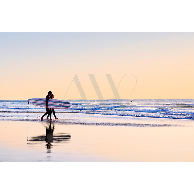 Two people walk along a serene beach at sunset, carrying a striped surfboard. The sky is a gradient of warm colors, reflecting on the wet sand and calm waves.