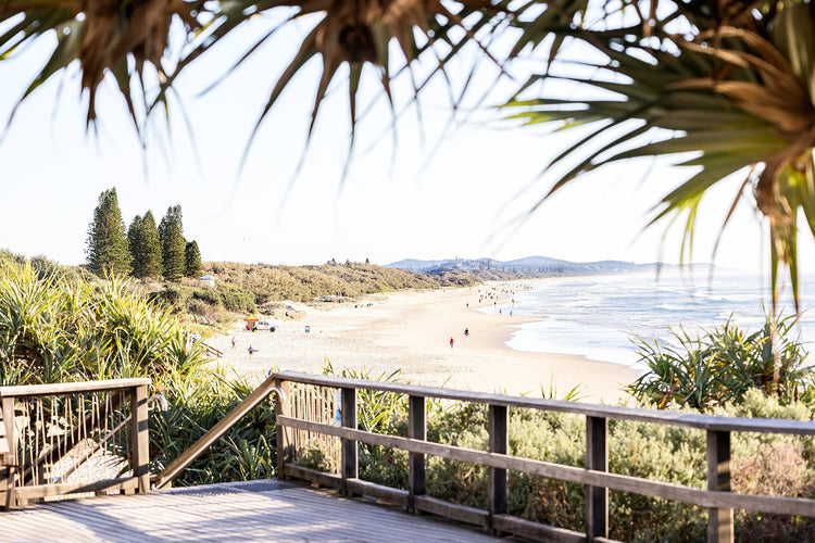 A sunny beach scene with a wooden boardwalk leading to golden sand and calm ocean waves. Pine trees and lush greenery frame the view. People can be seen enjoying the beach in the distance.