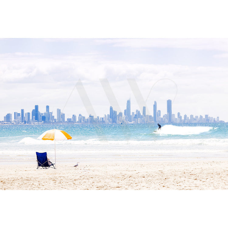 A beach with a yellow and white umbrella and a blue chair on the sand. A person surfs in the waves, and a city skyline looms in the background under a cloudy sky.