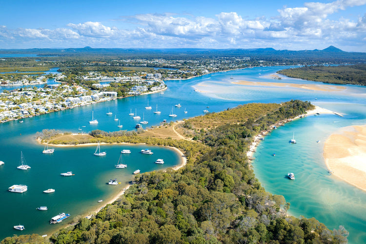 Aerial view of a coastal landscape with winding waterways, sailboats, sandy shores, and lush greenery. Residential areas are visible on the horizon, under a partly cloudy sky.