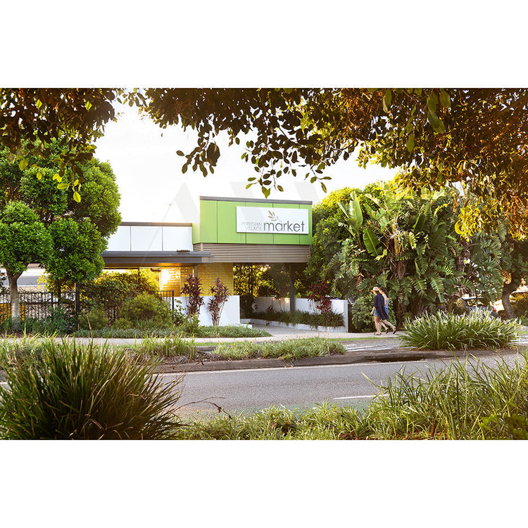 A modern building with a green sign reading market is seen through lush trees and plants. Two people walk on the sidewalk in front, and a road runs across the bottom of the image.