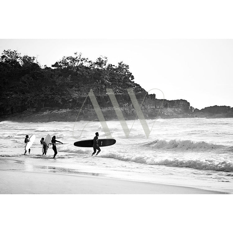 A group of surfers carrying surfboards head towards the ocean waves. The beach is sandy with a rocky hill in the background. The image is in black and white.