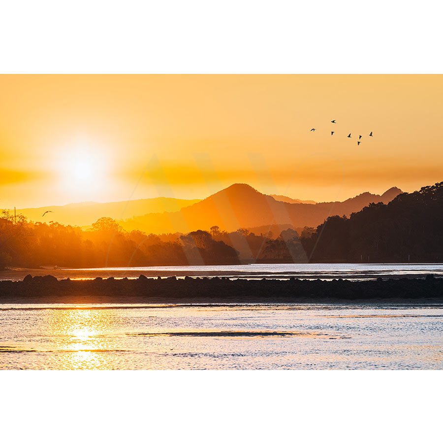 Brunswick Heads Image 8617 by AWP Image Library captures a serene sunrise over a calm lake, silhouetted mountains in the hinterland, and a gradient orange-yellow sky. Birds soar above the Brunswick River as vibrant colors reflect off the water.