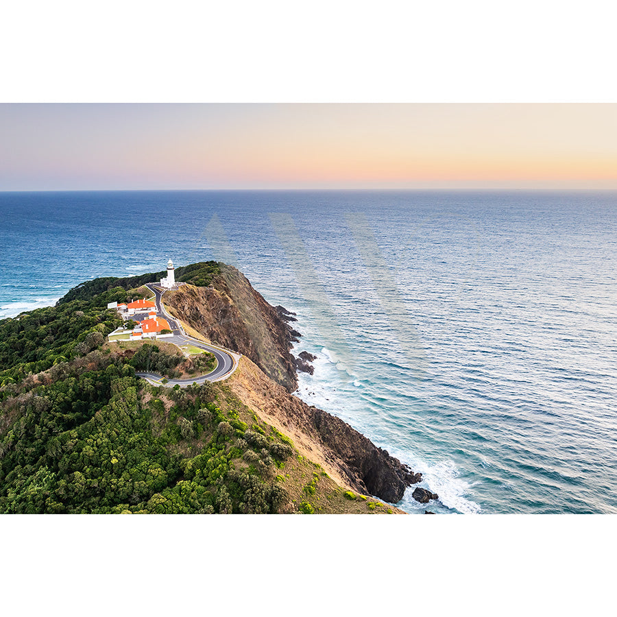 The Byron Bay Lighthouse Drone Image 0479 from AWP Image Library captures a coastal scene with a winding road to the iconic cliffside lighthouse, surrounded by lush greenery and ocean, under the clear warm light of sunrise or sunset.