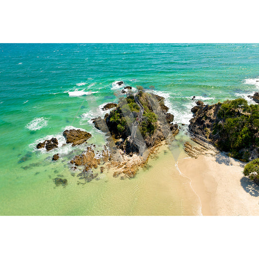The Byron Bay The Pass Drone Image 0441 by AWP Image Library showcases a rocky outcrop with turquoise waters, a staircase to a path, Wategos Beach lined with greenery, and waves gracefully hitting the rocks and shore.