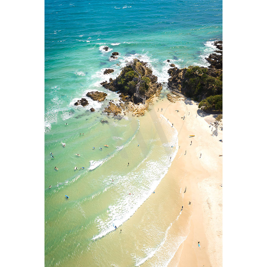 The Byron Bay The Pass Drone Image 0375 by AWP Image Library captures a stunning aerial view of a sandy beach with turquoise waves, scattered rocks, surfers, and beachgoers. Rocky outcrops extend into the ocean amidst clear, vibrant waters.
