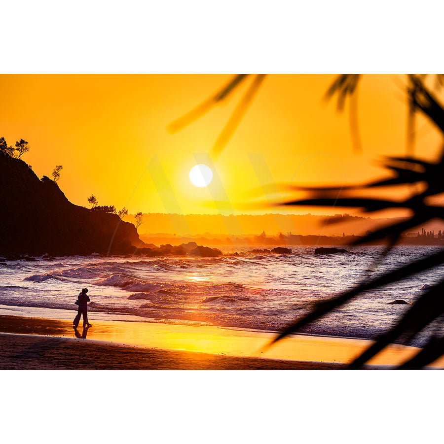 A silhouette of a person strolls along a sunset beach in Byron Bay Wategos Image 6999 by AWP Image Library, with the sun casting an orange glow over the waves. Trees and plants frame the scene and a rocky coastline appears on the horizon.