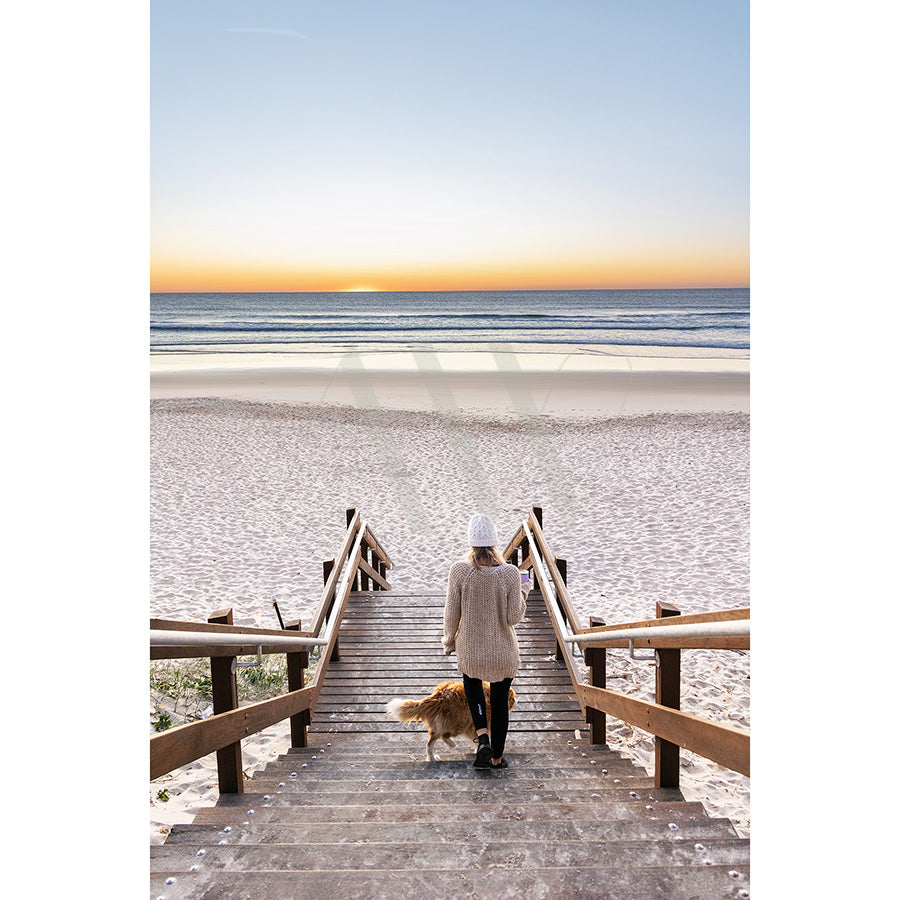 In Coolum Beach Image 8893 from AWP Image Library, a person in a sweater and beanie descends wooden stairs to a sandy beach with their dog, against the backdrop of an ocean and horizon visible under a clear sunset sky.