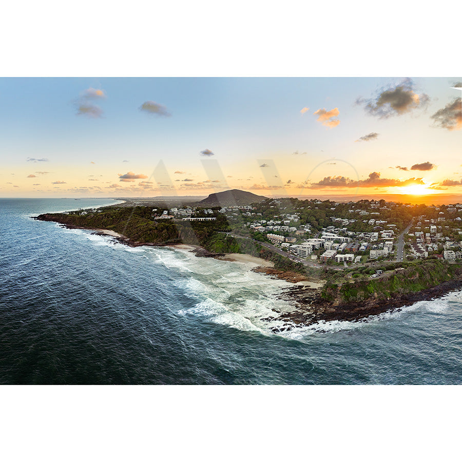 Aerial view captured in Coolum Beach Drone Image 0165 by AWP Image Library showcases a coastal town at sunset with rocky shores, houses on green hills, calm ocean, scattered clouds, and the sun setting on the horizon.