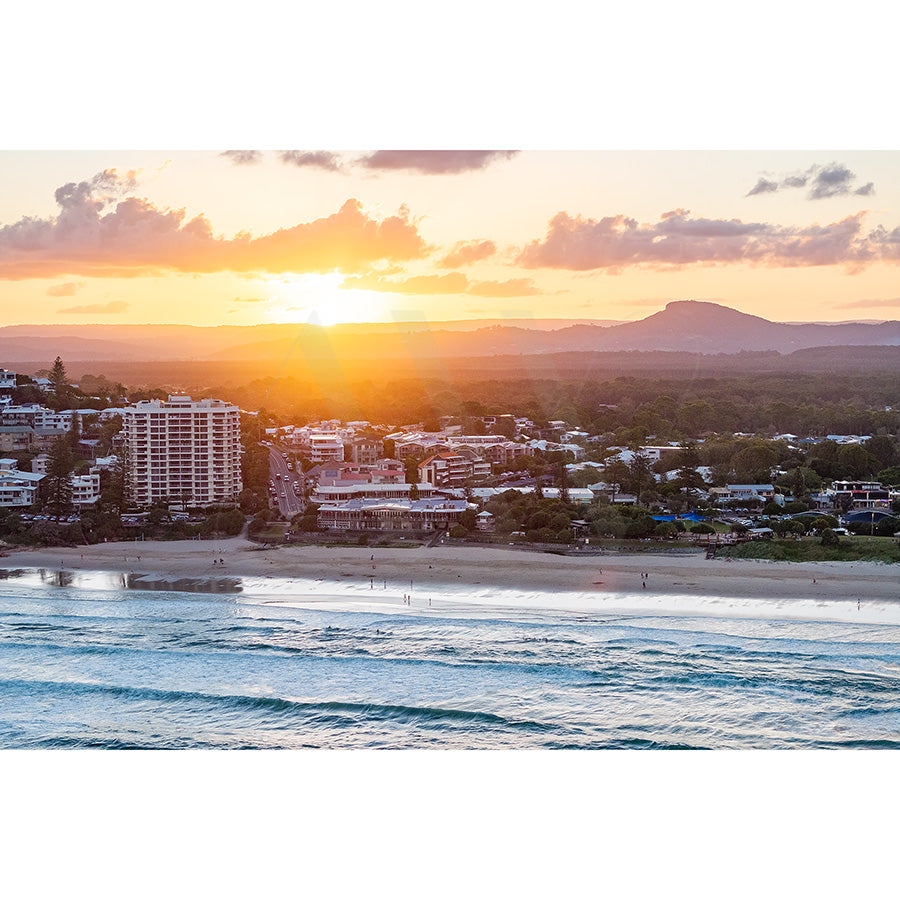 Coolum Beach drone Image 0190 by AWP Image Library showcases a coastal cityscape at sunset with gentle waves and a beach in the foreground, high-rise buildings and houses along the shore, and distant mountains under a partly cloudy sky.