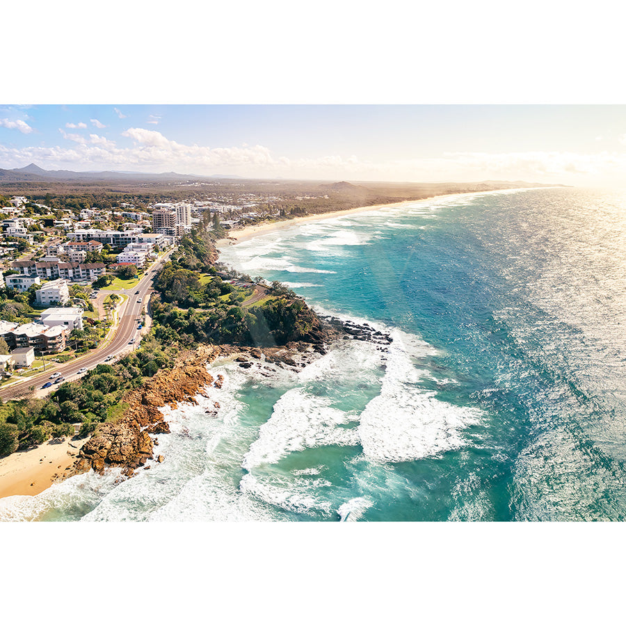The Coolum Beach Drone Image 0338 from AWP Image Library captures an aerial view of a coastal landscape with rocky cliffs, sandy beaches, turquoise waves, residential buildings, and a highway under a clear blue sky.