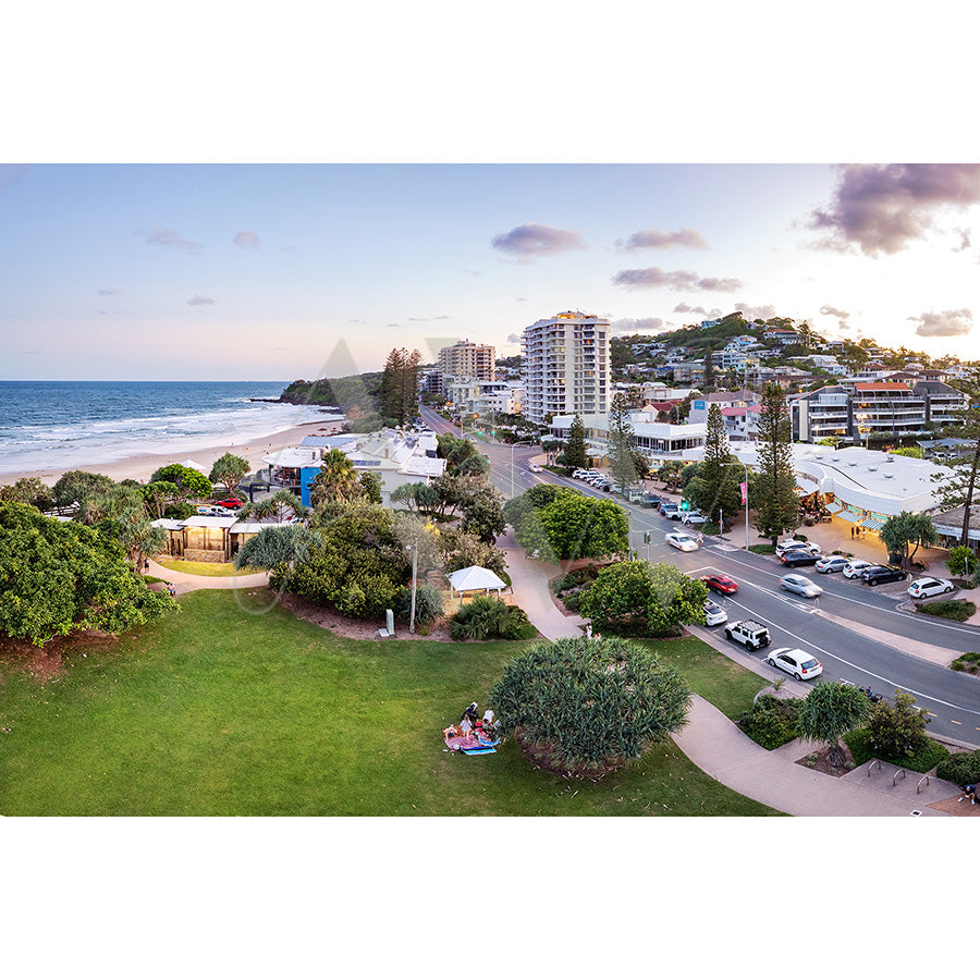 The Coolum Beach Drone Image 0448 from AWP Image Library captures a scenic coastal view of a seaside town with modern buildings, a lush green park, and a sandy beach. Cars are parked along the road as the ocean stretches into the distance under a partly cloudy sky.