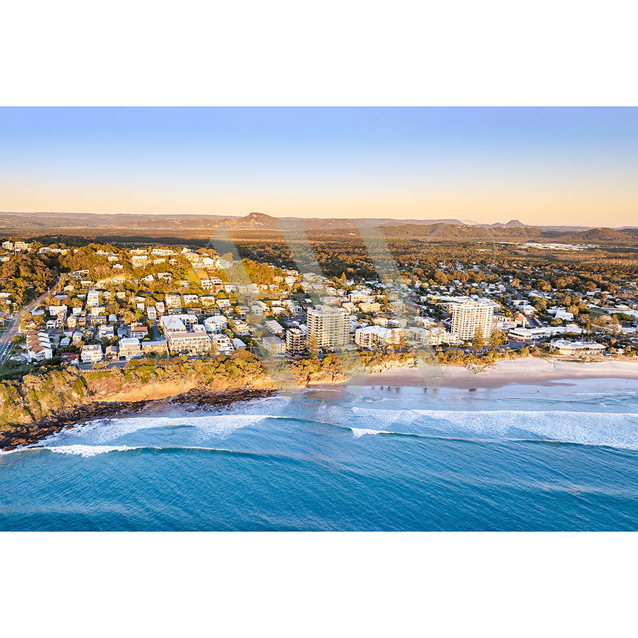 The Coolum Beach Drone Image 0629 by AWP Image Library showcases a charming coastal town with residential buildings and sandy shores. Ocean waves gently embrace the coastline under a clear, blue sunset sky, with hills and greenery creating a stunning backdrop.