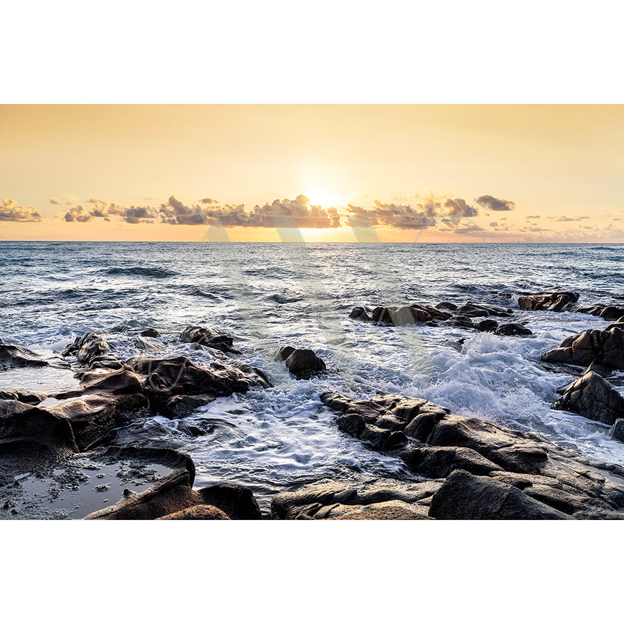 The Coolum Beach Image 0319 by AWP Image Library captures a serene coastal scene with waves crashing against rugged rocks, a warm golden sun on the horizon, and fluffy clouds dotting the sky.