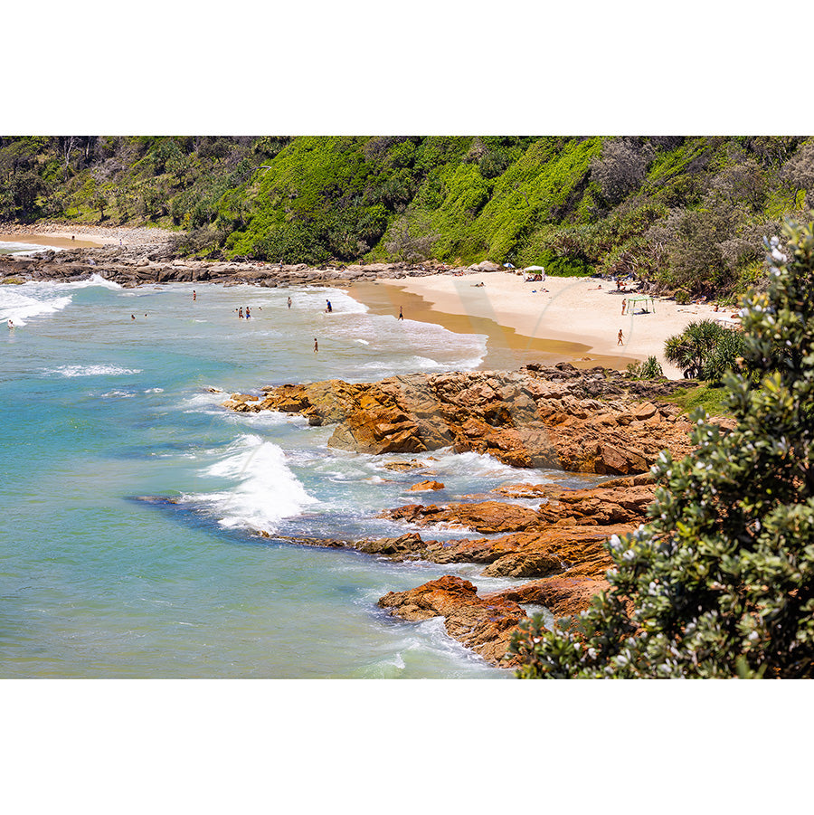 The Coolum Beach Image 0445 from AWP Image Library showcases a scenic view of a coastal beach with turquoise waves crashing against rocky shores, people relaxing and swimming on the sandy beach, lush green vegetation in the background, all under a sunny and vibrant atmosphere.