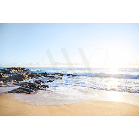 The Coolum Beach Image 0479 from AWP Image Library captures a serene beach with golden sand, gentle waves, and scattered rocks under a clear blue sky while sunlight reflects on the water.