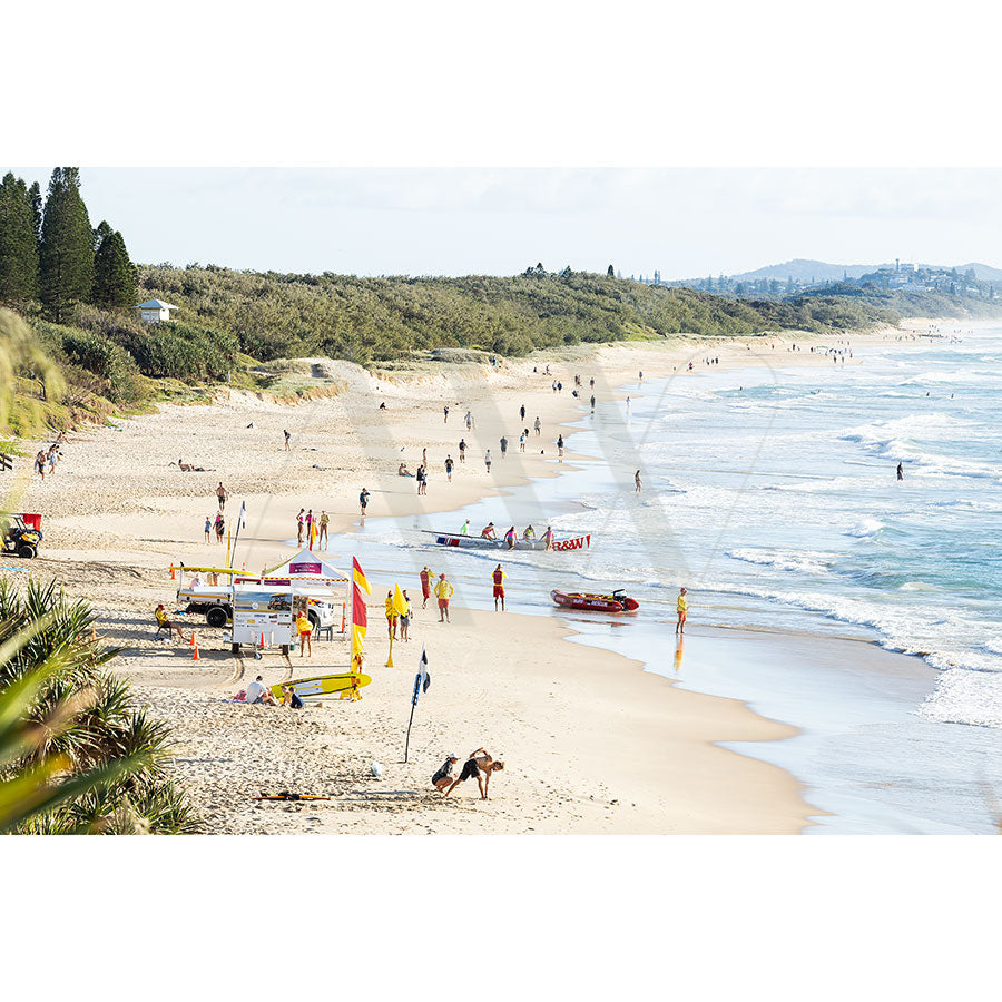 The Coolum Beach Image 0539 from AWP Image Library captures a beach scene with people enjoying the sand and surf, lifeguards in yellow and red uniforms near a patrol tent and rescue boat, extended coastline, greenery on dunes, and crashing waves.