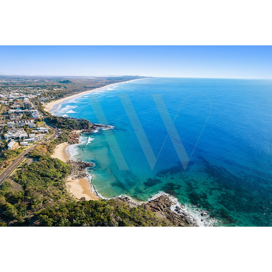 Coolum Beach Image 0753 from AWP Image Library captures an aerial view of a coastal landscape with a sandy beach, vibrant turquoise ocean, and lush greenery. Buildings nestle near the shoreline under a clear blue sky stretching to the horizon.