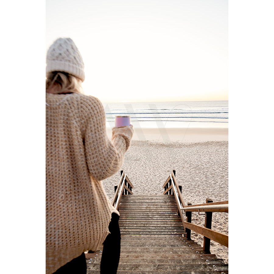 In the Coolum Beach Image 8892 by AWP Image Library, a person in a knitted sweater and beanie walks down wooden steps towards a sandy beach, holding a cup. The ocean is visible under a clear sky in the distance.