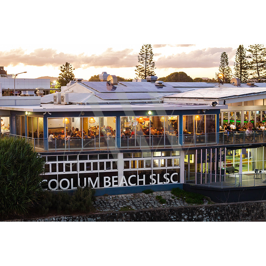 A vibrant beachfront scene with large windows and a bustling crowd inside, featuring COOLUM BEACH SLSC on the outside. The overcast sky and trees in the background add to the warm, inviting atmosphere. Captured in Coolum Beach Surf Club Image 0620 from AWP Image Library.