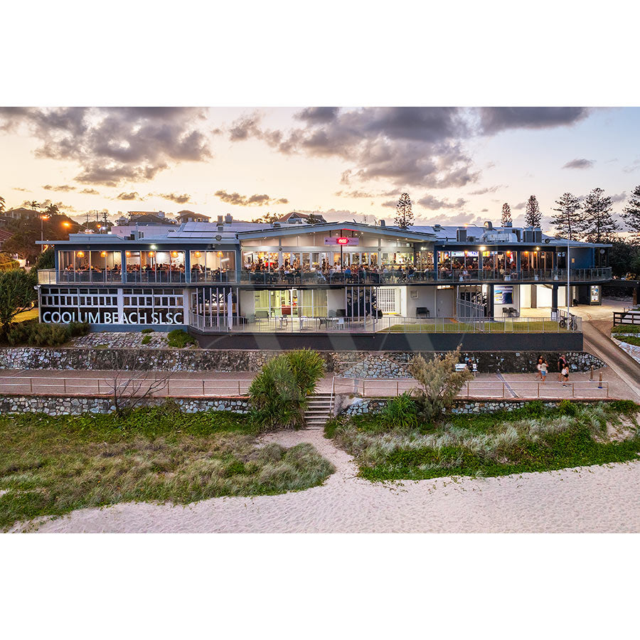 The Coolum Beach Surf Club Image 0645 from AWP Image Library features a modern two-story building with large glass windows and an ocean view by the sandy beach. The scene includes people inside, a partly cloudy sky, greenery, and a walking path leading to the entrance.