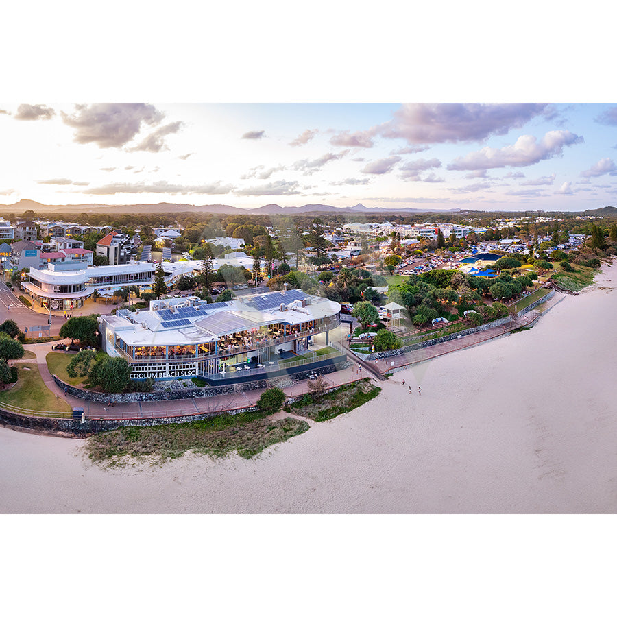 The Coolum Beach Drone Image 0563 by AWP Image Library captures a coastal town at sunset, showcasing modern buildings and a shoreline walkway with few people near Coolum Beach. The partly cloudy sky casts a warm glow over the scene, just beyond the renowned Coolum Surf Club.
