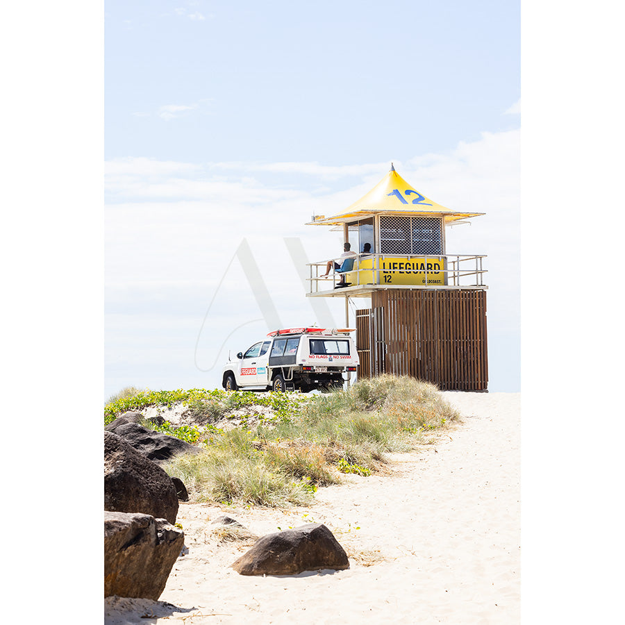 Currumbin Beach Image 4444 by AWP Image Library captures a lifeguard tower numbered 12 on a sandy beach, with a parked lifeguard vehicle, under vibrant sunshine. The foreground features lush greenery and rocks beneath a clear blue sky.