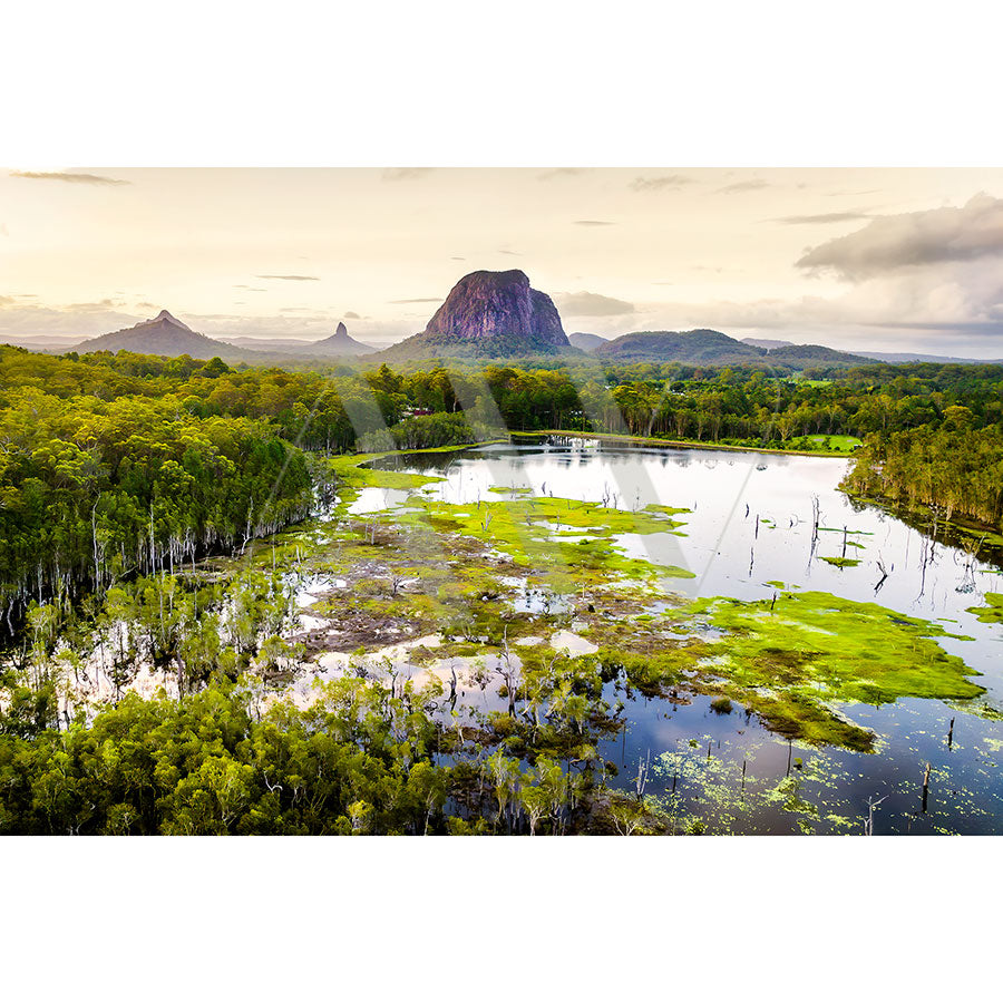 The Glass House Mountains Drone 0481 from AWP Image Library captures an aerial view of a lush landscape featuring a large lake surrounded by greenery, with a distinctive rocky mountain in the background under cloudy skies. Patches of moss or algae add texture to the water.
