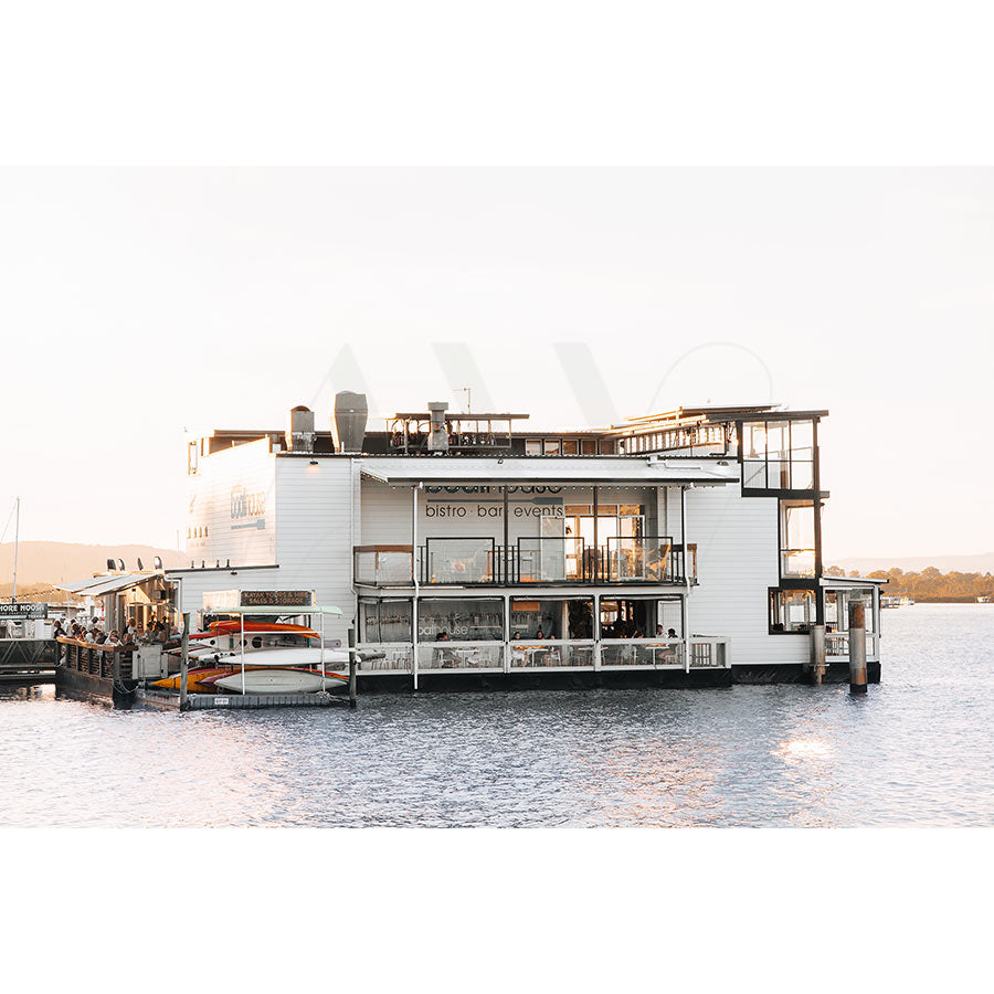 The Gympie Tce Boathouse by AWP Image Library floats on the Noosa River, resembling a contemporary Boat House with large windows and multiple decks. Its signage reads bistro bar events, amidst calm waters and clear skies, with distant land and occasional seagull calls.