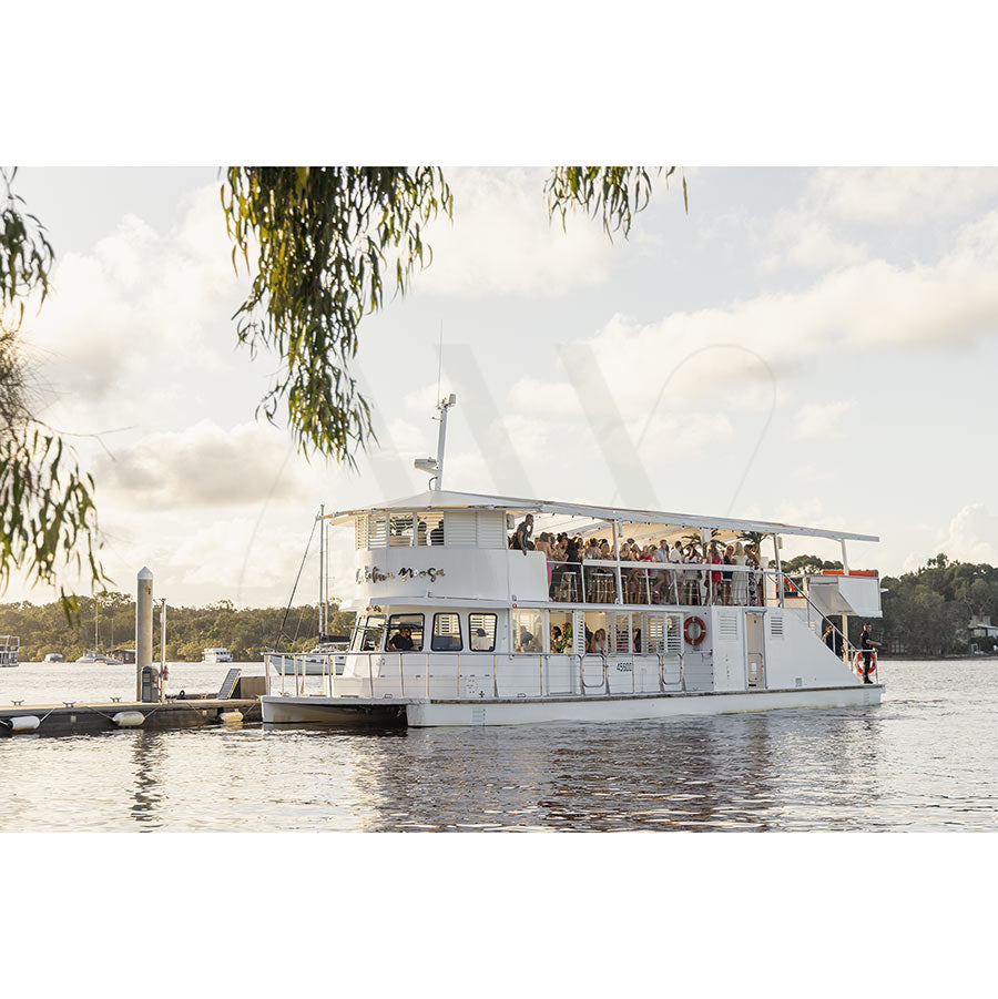 Gympie Tce Image 3358 by AWP Image Library features a white, two-level riverboat cruising near a dock on a calm river with trees and clouds, as guests savor gourmet canapés onboard.