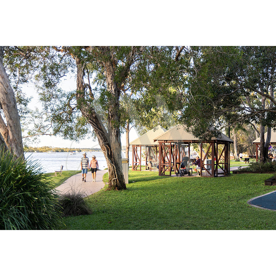 The Gympie Tce Image 3407 from AWP Image Library shows people relaxing on green lawns under large trees, with two walking along accessible riverside pathways and picnic shelters by the Noosa River foreshore.