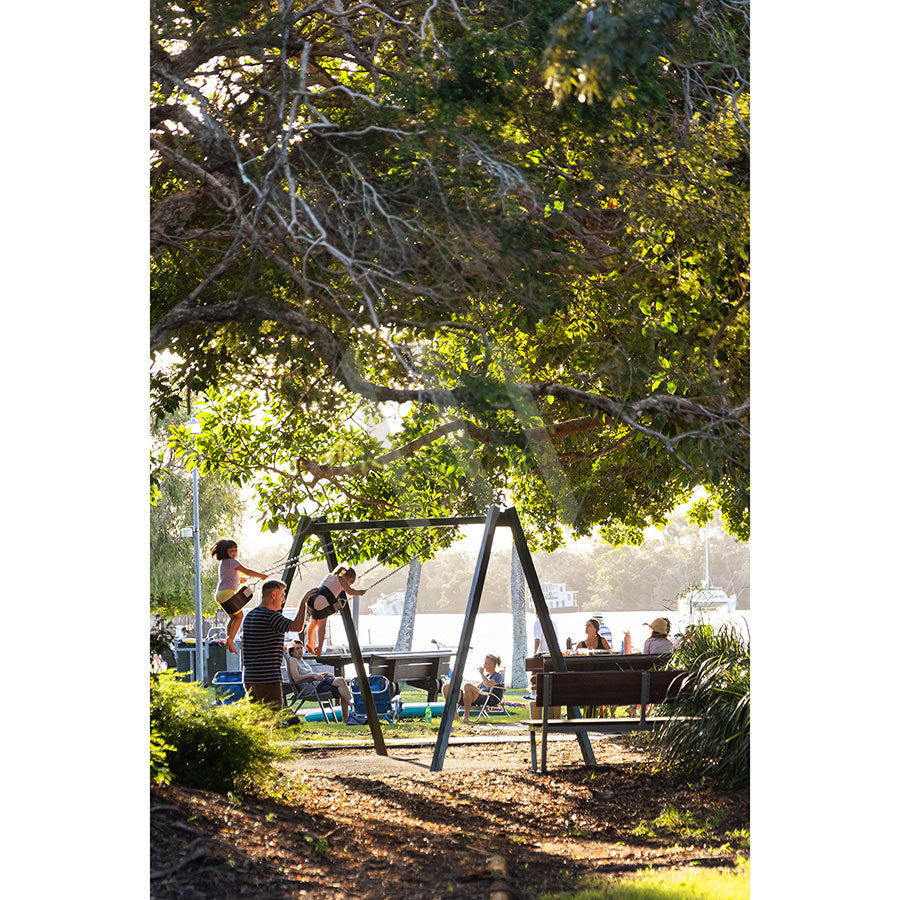 In Gympie Tce Image 3424 from AWP Image Library, children play on a swing set beneath leafy trees while adults relax on benches by the Noosa River, enjoying the sunlit, laid-back atmosphere.