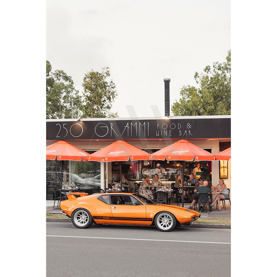 Gympie Tce Image 3595 by AWP Image Library features an orange vintage sports car parked near boutiques and a food & wine bar, with people dining outside under red umbrellas on a cloudy day.