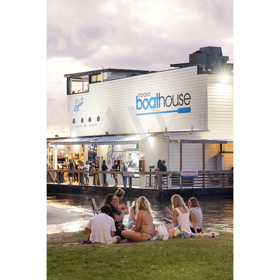 A group relaxes on the grass by the water at sunset with Noosa Boathouse in the background, lit and lively. Photo: Gympie Tce Image 3663, AWP Image Library.
