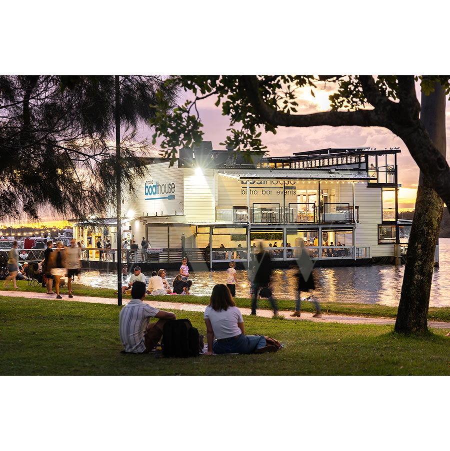 At sunset, people relax on the grassy riverfront by the water as The Boathouse Noosa glows and live music drifts nearby in AWP Image Library’s Gympie Tce Image 3716.