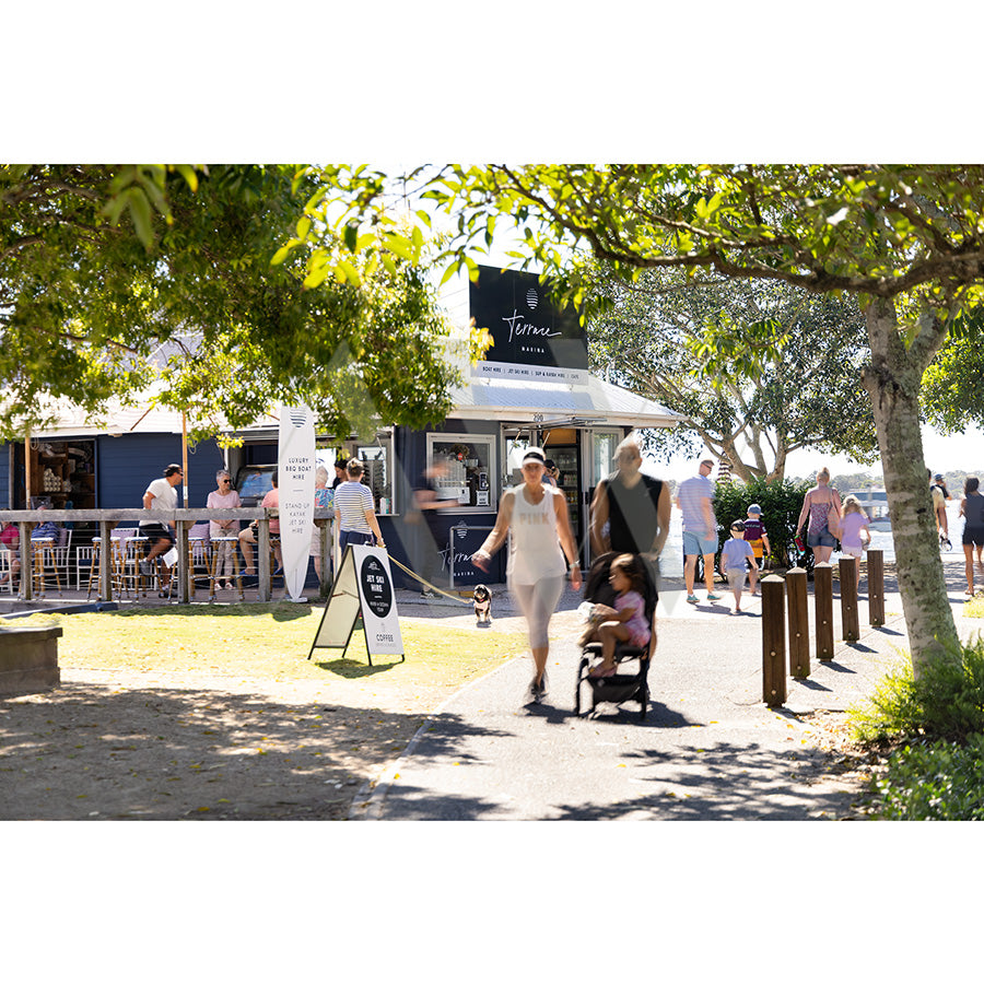In Gympie Tce Image 4041 by AWP Image Library, people enjoy a sunny day on Gympie Terrace—some relaxing at café tables, others strolling along Noosa foreshore, as a woman pushes a stroller amid trees and greenery.