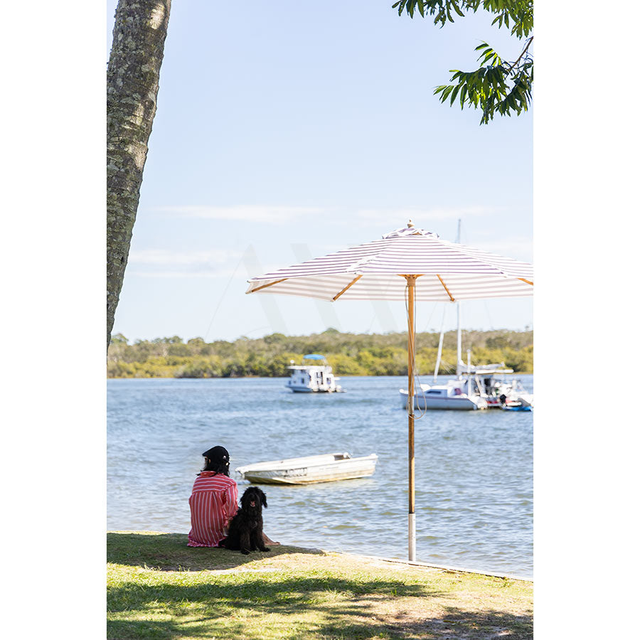 Enjoy outdoor living on a sunny Noosa day in Gympie Tce Image 3963 by AWP Image Library, featuring a person relaxing with their small black dog under a white umbrella by the water at Gympie Terrace, with boats and trees in the background.