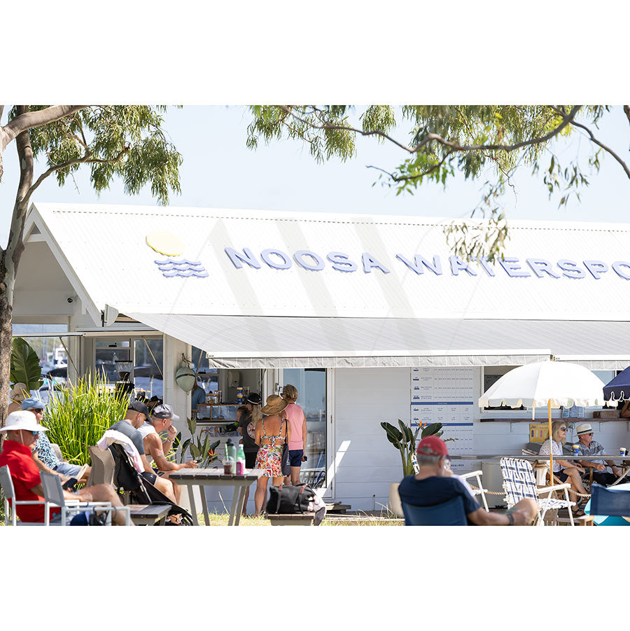 People relax and dine outdoors under umbrellas near a white building signed Noosa Watersports in Gympie Tce Image 4167 from the AWP Image Library, surrounded by greenery on a sunny day by the Noosa foreshore.