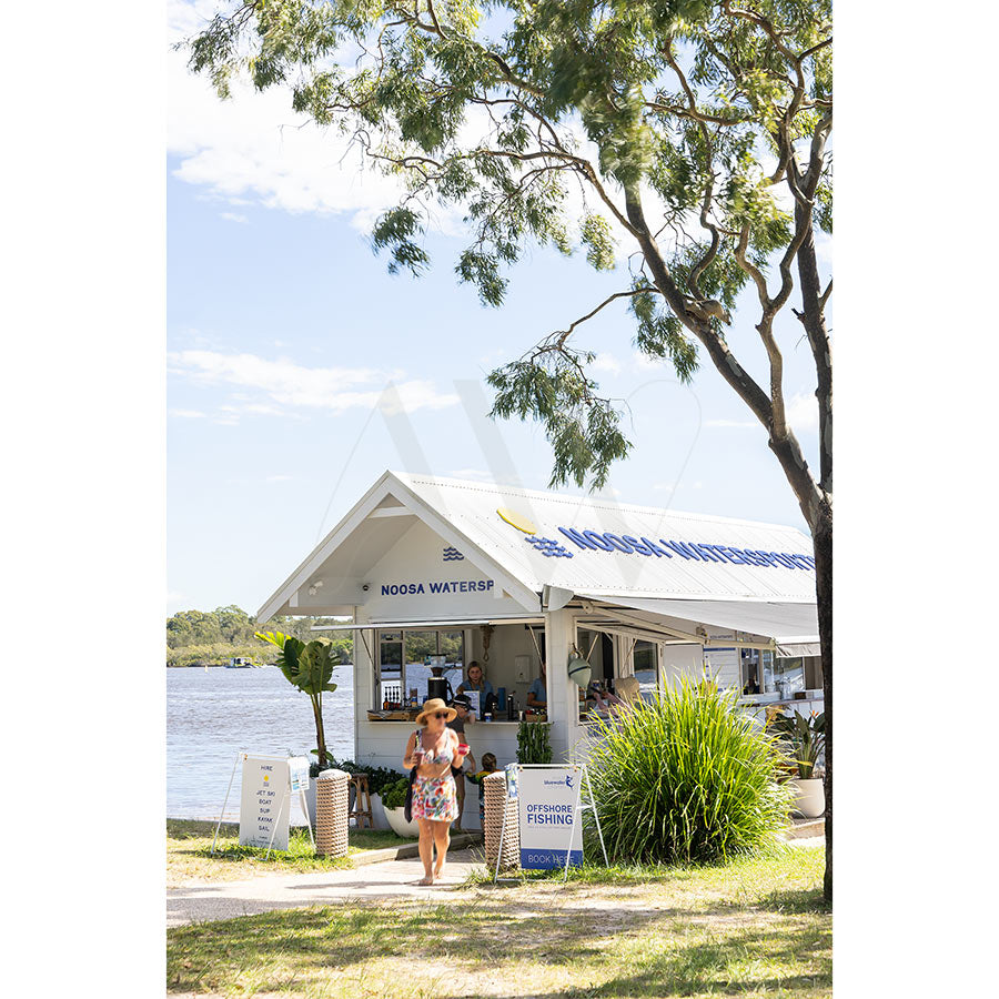Gympie Tce Image 4285 from AWP Image Library shows a small white riverside watersports rental shack on Gympie Terrace, with a woman in a hat and floral dress out front, surrounded by green trees under a sunny blue sky.