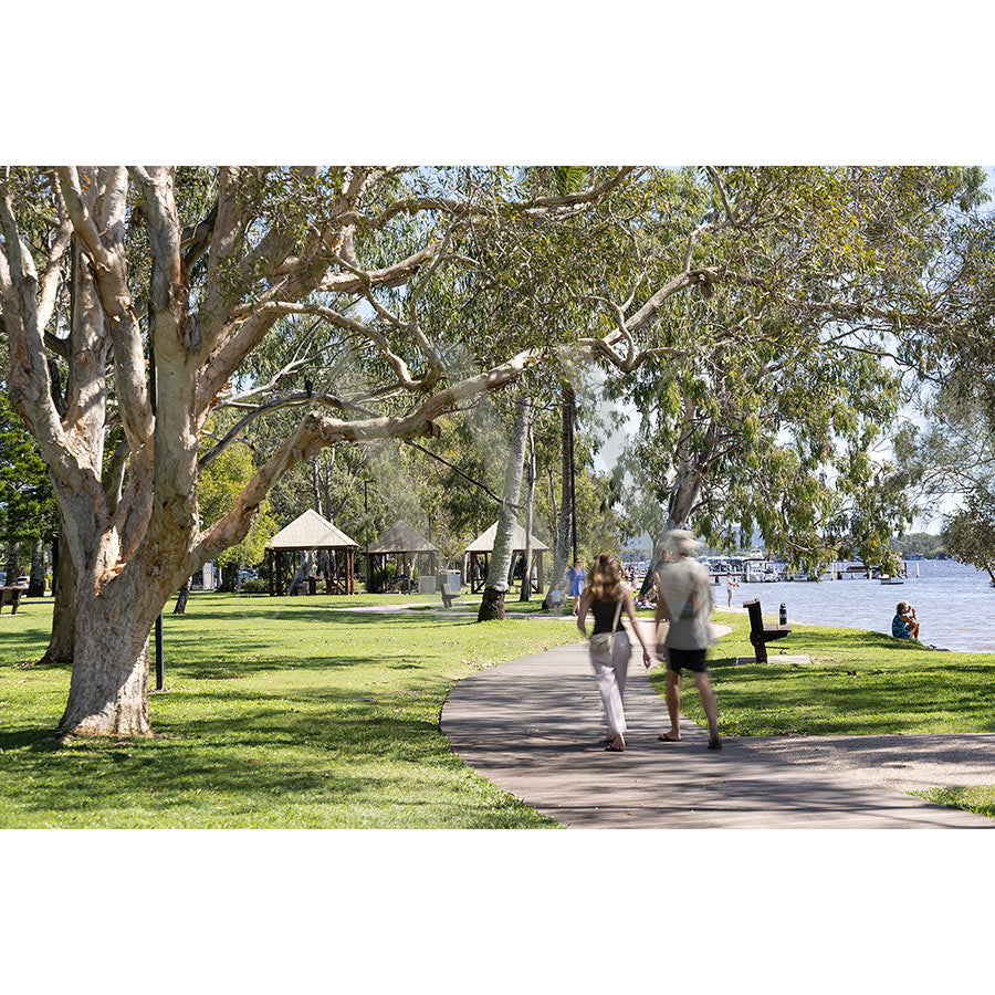 Gympie Tce Image 4752 by AWP Image Library captures a paved path winding through a leafy park with picnic shelters and walkers, while people relax along Noosa’s sparkling river foreshore under the sun.