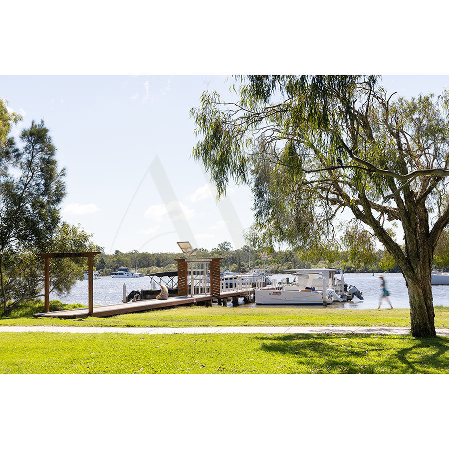 The Gympie Tce Image 4791 by AWP Image Library captures a peaceful riverside with boats, a dock, green grass, leafy trees, and a person strolling under a bright blue sky with clouds.