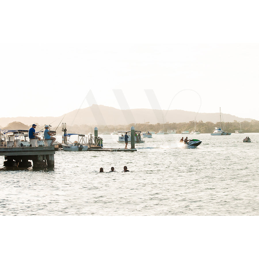 Gympie Tce Image 5830 by AWP Image Library captures a lakeside scene with boats and jet skis. Two people swim near a fishing pier, with mountains in the hazy background under a clear sky.