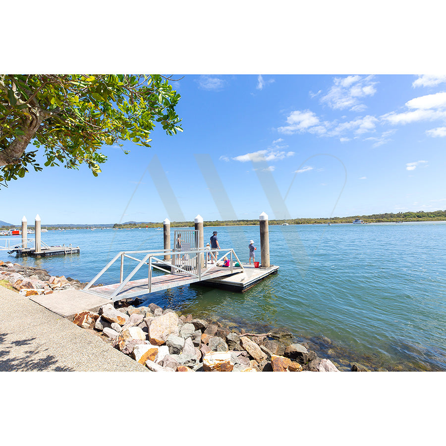 The AWP Image Librarys Gympie Tce Image 5881 captures two holidaymakers on a dock by the Noosa River, surrounded by clear blue water, a partly cloudy sky, rocky shores, and a tree providing shade. Another dock in the distance enhances the serene landscape.