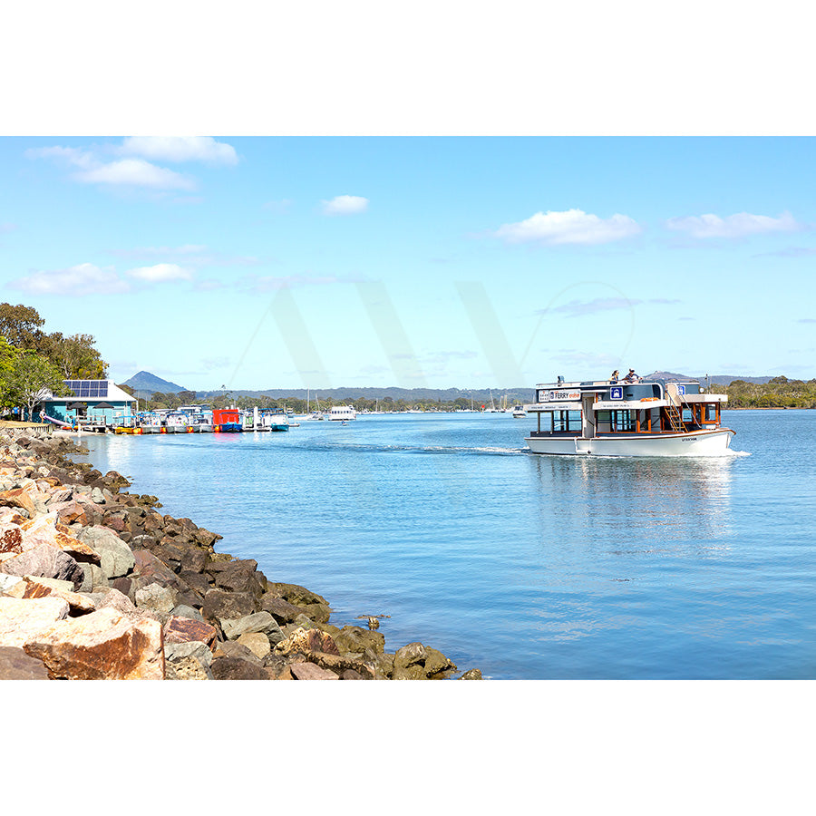 The Gympie Tce Image 5922 from AWP Image Library captures a scenic river with a ferry on clear blue waters. The riverbank features rocks and greenery, while several boats are docked in the distance under a bright sky with clouds.