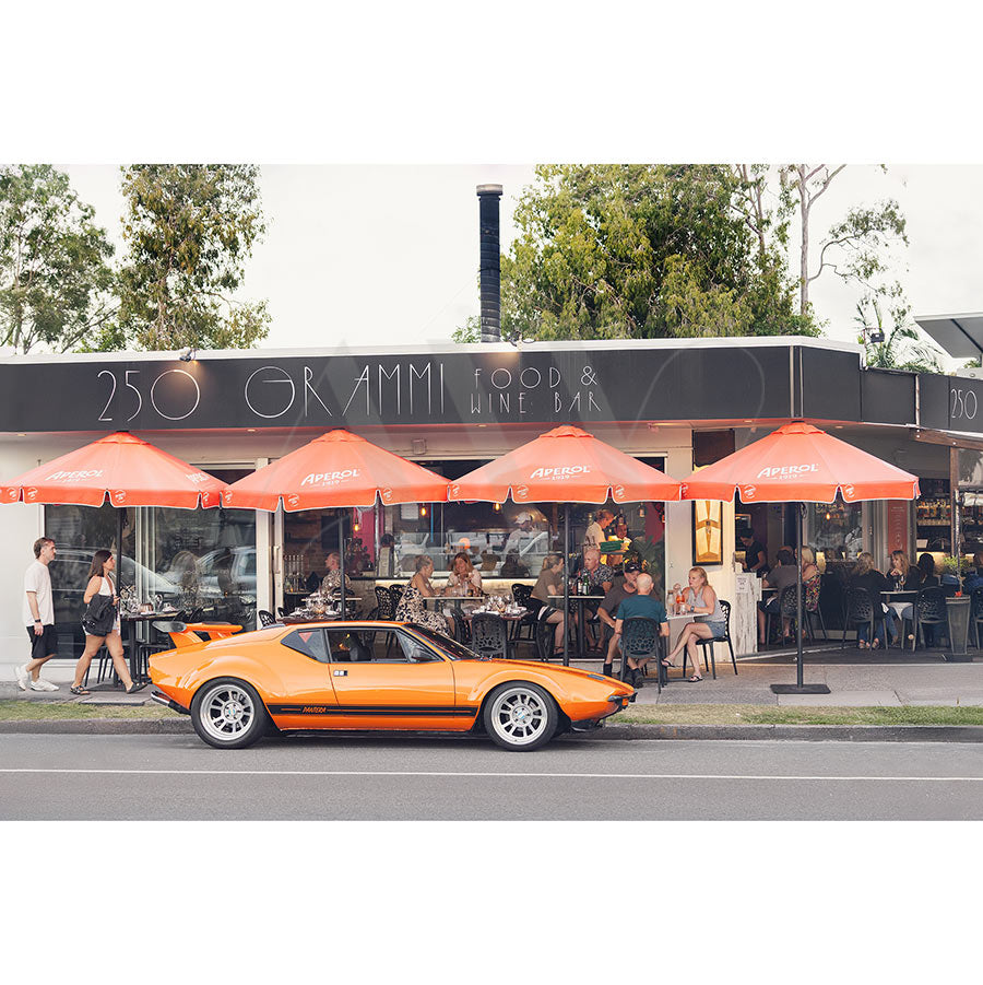 Gympie Tce Image 3589 by AWP Image Library shows an orange vintage sports car parked outside 250 Grammi Food & Wine Bar in Noosaville, where people enjoy wood-fired pizzas under bright orange umbrellas.