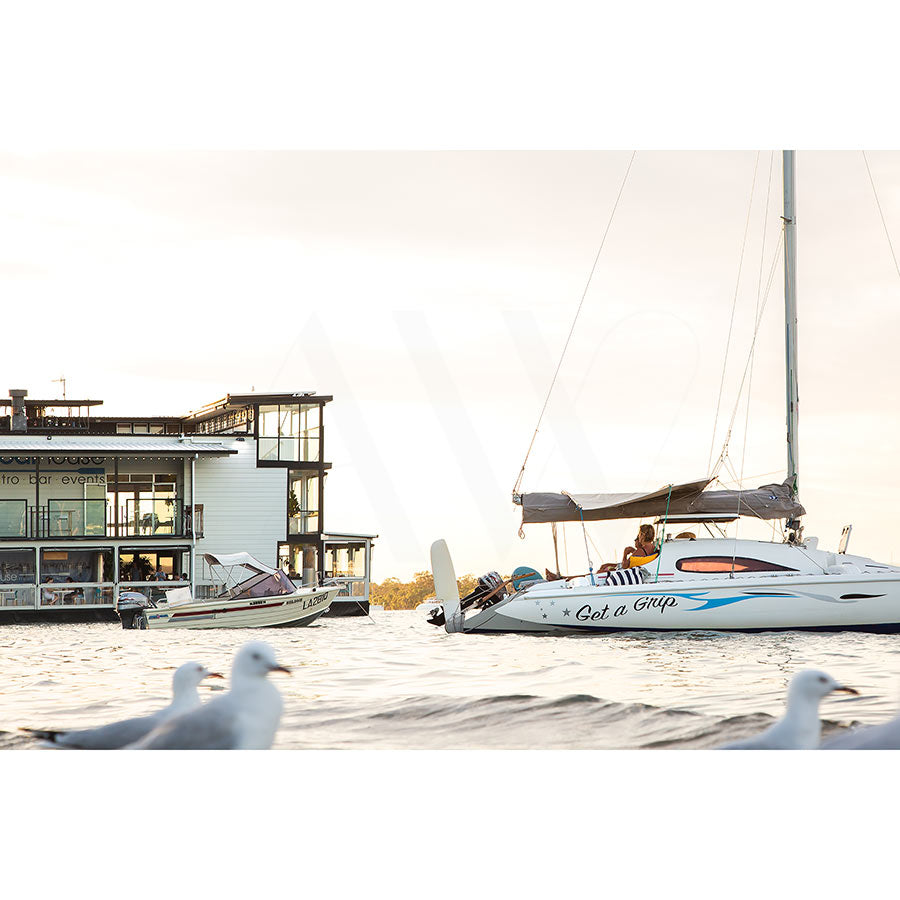 In Gympie Tce Image 8032 from AWP Image Library, the sailboat Get a Grip glides on Noosa River with passengers aboard. To the left, a white multi-story house stands gracefully; in the foreground, seagulls drift under an overcast sky, casting soft light over this tranquil scene.