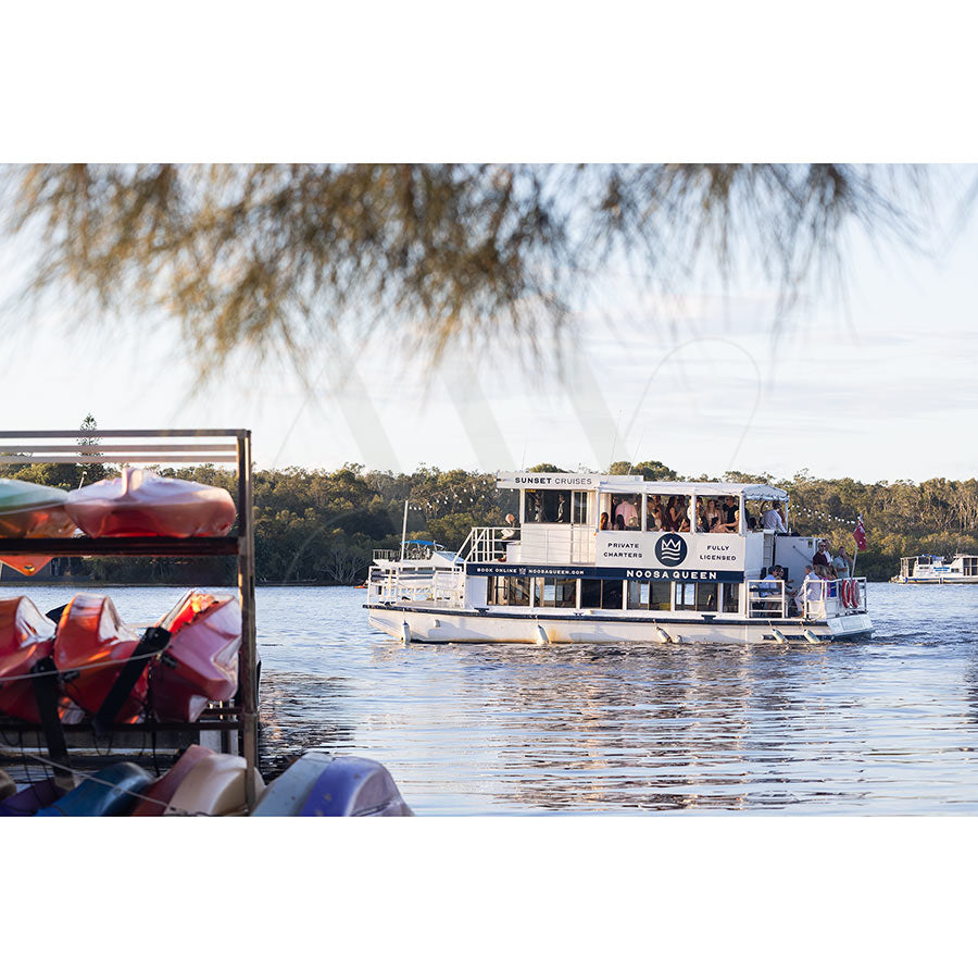 Gympie Tce Image 3492 by AWP Image Library captures a riverboat cruising on calm water, colorful kayaks stacked in the foreground, and trees in the background—a perfect evening scene under partly cloudy skies.