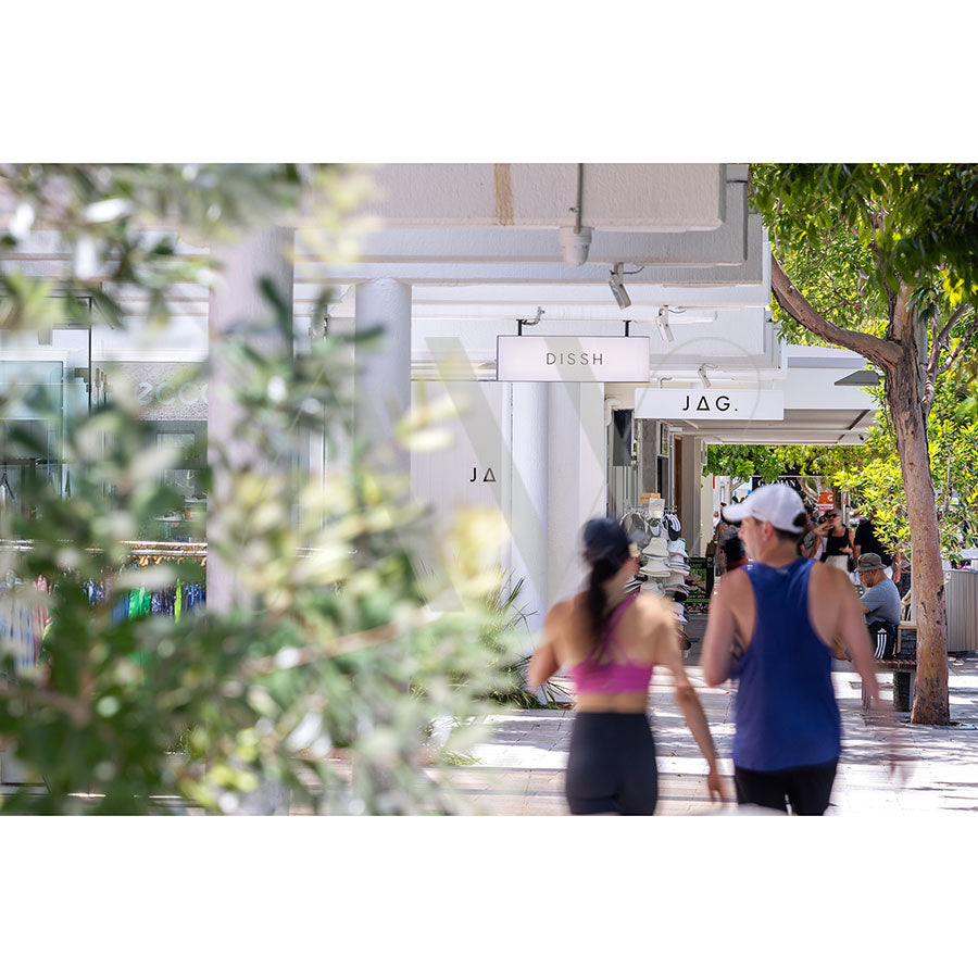 The Hastings Street Image 5370 from AWP Image Library captures two individuals in athletic wear strolling through a sunny, tree-lined shopping area with visible DISH and JAG store signs, enhanced by lush greenery.